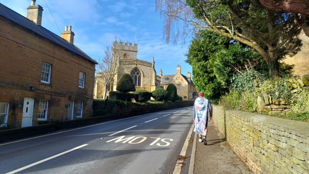 Dave Hollingsworth striding ahead on his fundraising challenge. He is walking towards a picturesque church in a small village.
