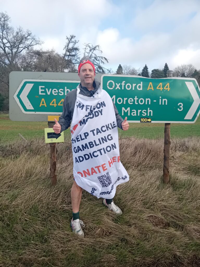 Dave Hollingsworth standing in front of a road sign that points to Evesham in one direction and Moreton-in-Marsh in the other direction. He is wearing his Gordon Moody fundraising flag detailing his challenge and how to donate.