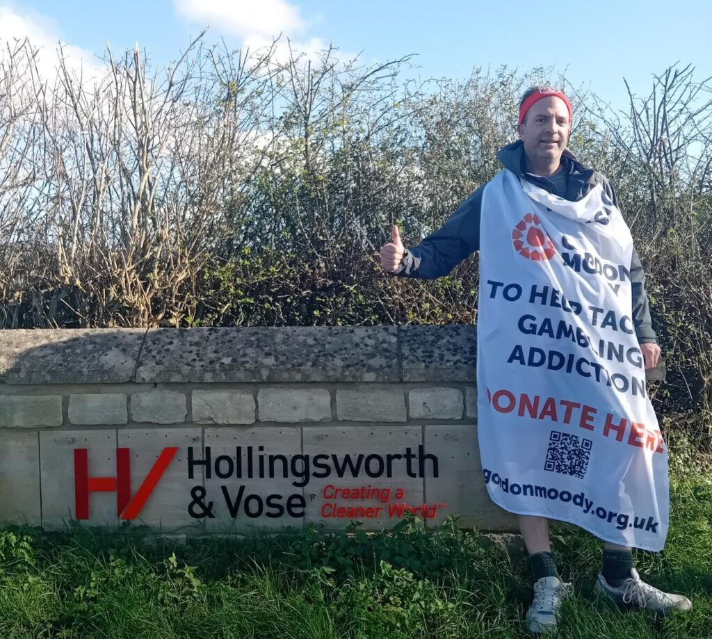 Dave Hollingworth standing beside a sign for the Hollingsworth & Vose paper mill in Winchcombe, near Cheltenham. Dave is wearing a Gordon Moody flag with the details of his fundraising challenge.