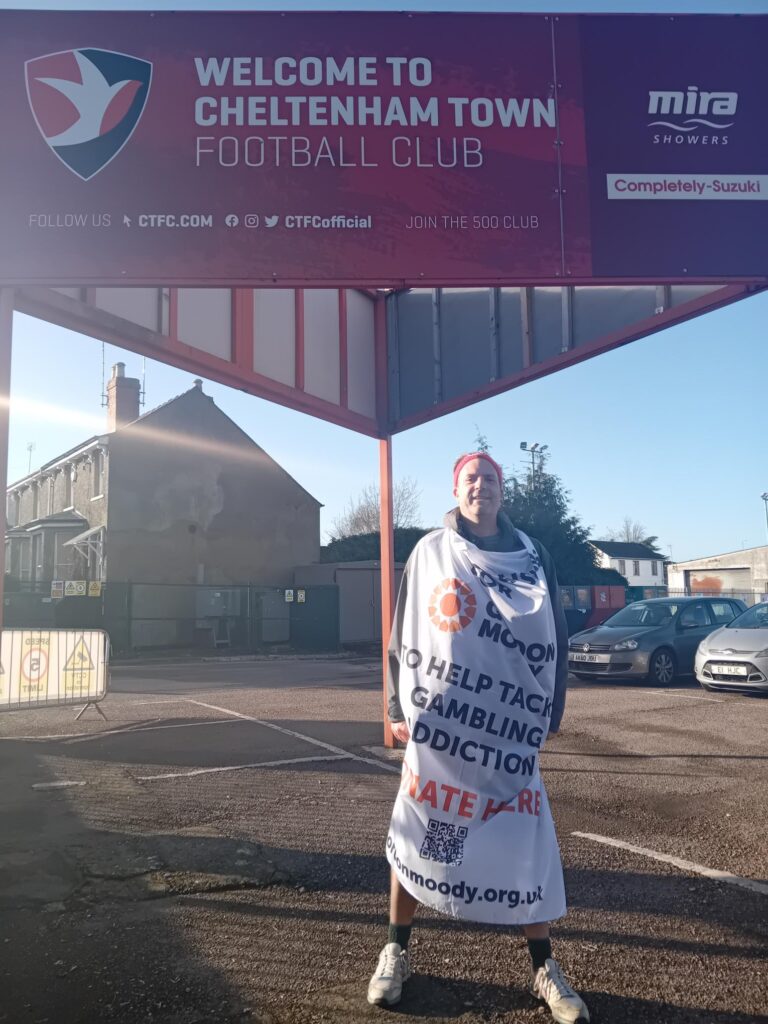 Dave Hollingsworth standing underneath a Welcome to Cheltenham Town FC sign, whilst wearing a flag containing details of his fundraising walk for Gordon Moody.