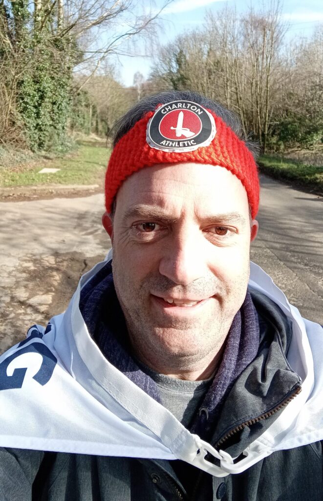 Dave Hollingsworth smiling towards the camera whilst on his fundraising walk. He is wearing a Charlton Athletic FC beanie hat and a Gordon Moody flag detailing his challenge.