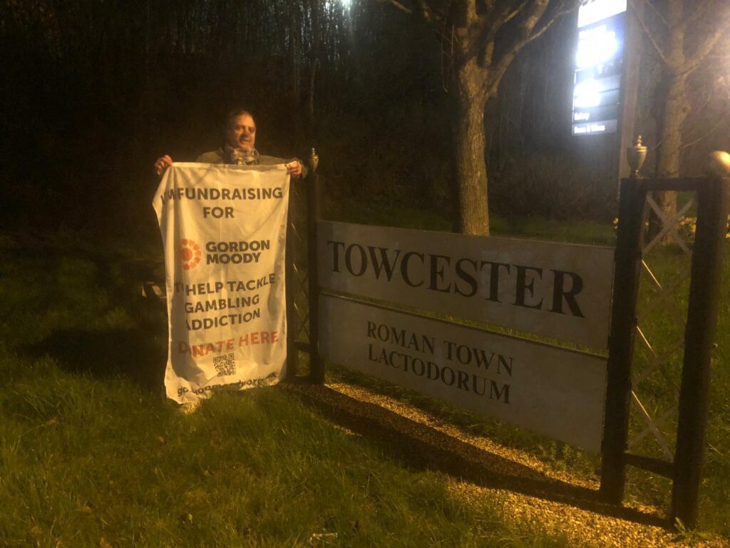 Dave Hollingsworth standing beside a sign reading Towcester Roman Town Lactodorum. Dave is holding a large white flag with the details of his fundraising challenge.