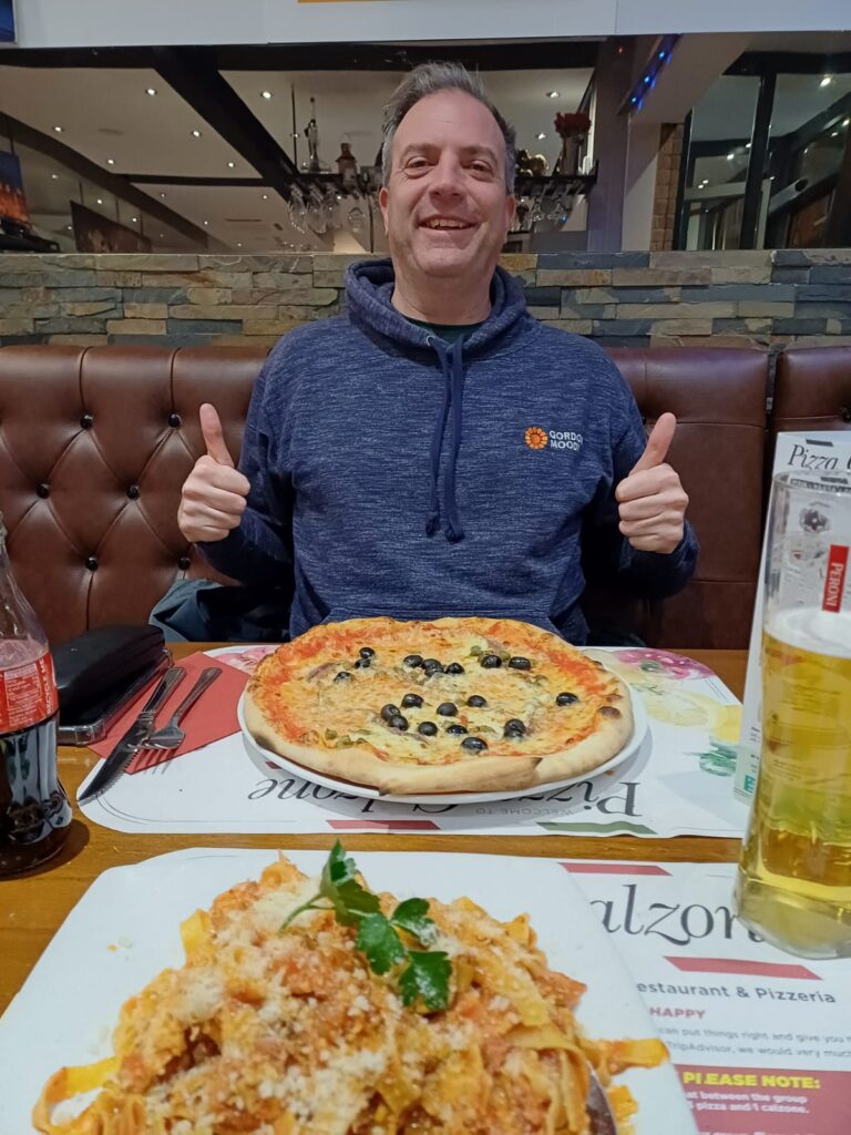 Image of Dave Hollingsworth wearing a Gordon Moody branded hoodie holding up two thumbs in an Italian restaurant with a pizza with black olives in front of him and a plate of pasta in front of the camera man.