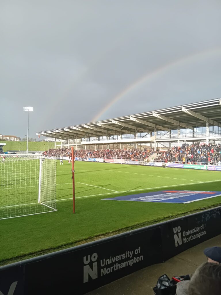 The view from behind one of the goals at the Charlton Athletic FC game away at Northampton Town. Behind one of the stands is a rainbow in a sky of blue.
