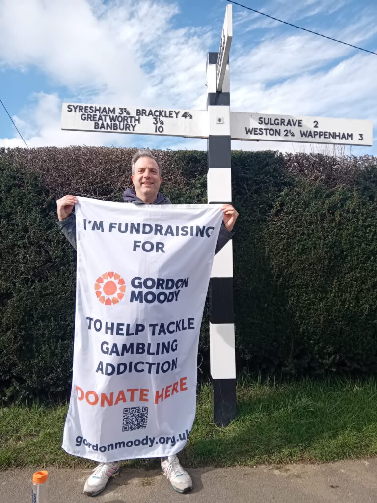Dave Hollingsworth standing in front of a finger post road sign whilst holding his Gordon Moody fundraising flag showing how to donate to his challenge.