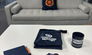 Selection of Gordon Moody branded items - notebook, pen, mug, lunch bag, and lanyard - on a white desk with a grey sofa in the background with a Gordon Moody branded cushion on it.