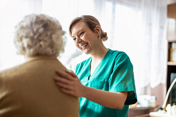 A caring healthcare professional smiling as she attends to an elderly patient in a warm, sunlit room.