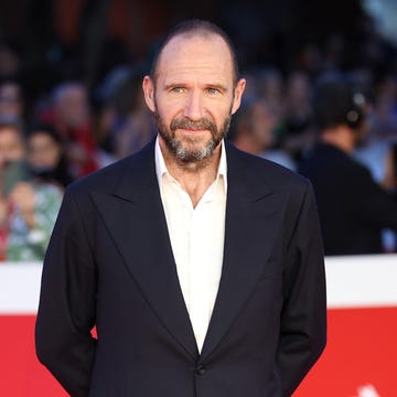 ralph fiennes on a red carpet, dressed in a dark suit and white shirt, the background features a crowd of onlookers