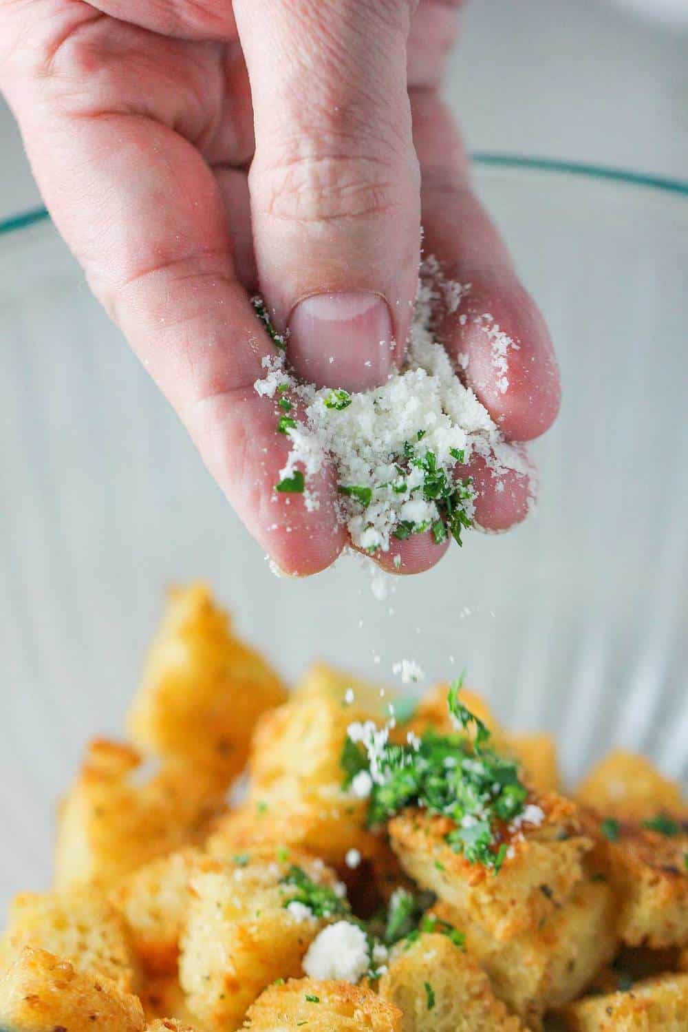 A hand sprinkling cheese and herbs onto baked croutons.