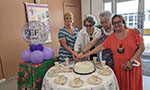 Quatro mulheres cortando um bolo sobre uma mesa. Elas estão com o braço direito estendidos em direção ao bolo como se fossem cortar juntas. Expressão facial feliz ou neutra. No lado da mesa, um balão com o texto “REF 30 anos” e um banner de divulgação.