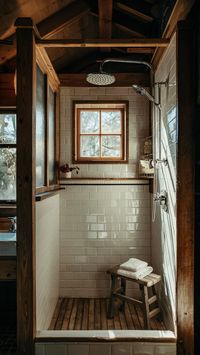 Discover the charm of this rustic bathroom featuring a wooden-framed shower enclosure with white tiles and a stainless steel showerhead. A wooden bench with a folded towel adds a cozy touch, while natural light filters through a small wooden-framed window, illuminating the wooden beams and tiled floor.