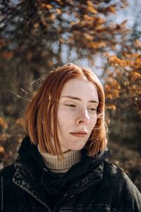 A calm pretty girl with red hair in front of the trees.