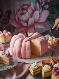 A photograph of a pink pumpkin shaped cake being cut with a sharp knife, surrounded by a flowery girly dimmed background