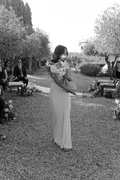 a woman in a long white dress walking down the aisle with flowers on her hand