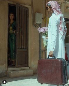 a man with a suit case and flowers in front of a door, holding a suitcase