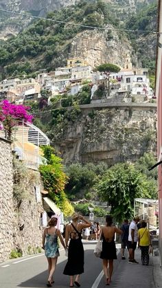 several people walking down a narrow street in front of some buildings and mountains with houses on the hill behind them