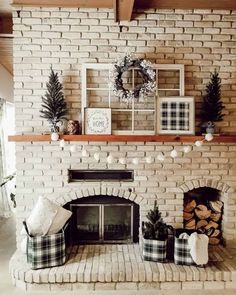 An earth-toned brick fireplace with a wood mantle is simply decorated with miniature potted fir trees, a hollow wood window frame, a wreath with pearl sprays, and green plaid accents. A white pom-pom garland drapes over plaid linen baskets beside the fireplace and wood storage...   Image: ourhappilyeverhome