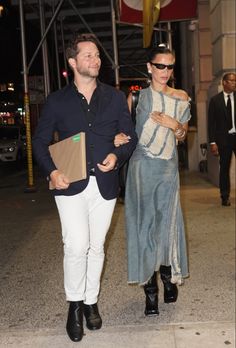 a man and woman are walking down the street at night, dressed in white pants