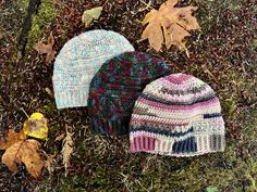 three knitted hats laying on the ground next to fallen leaves and dead grass,