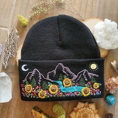 a black beanie hat with sunflowers and mountains on it next to other items