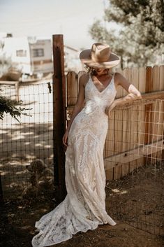 a woman in a white dress and cowboy hat leaning against a fence with her back to the camera