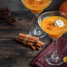 two glasses filled with orange juice and cinnamon on top of a wooden table next to pumpkins