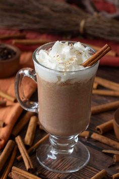 a glass mug filled with hot chocolate and whipped cream surrounded by cinnamon sticks on a wooden table