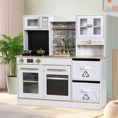 a toy kitchen with white cabinets and black appliances on the floor in front of a potted plant