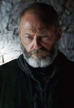 an older man with a beard stares into the camera while standing in front of a stone wall