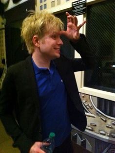 a man in a black jacket and blue shirt standing next to a window with his hand up