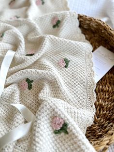 a knitted blanket with pink roses on it next to a wicker basket and white ribbon