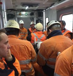 several men in orange jackets and hard hats on a boat or ship looking out the window