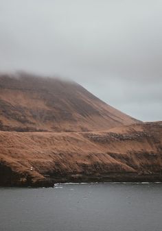 a large hill covered in brown grass next to a body of water on a foggy day