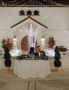 a bride and groom sitting at a table in front of a fountain with water shooting from it