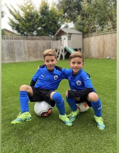 two young boys sitting on top of a soccer ball wearing blue and black uniforms with their arms around each other