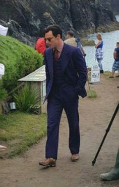 a man in a blue suit and sunglasses standing next to a body of water with other people around him