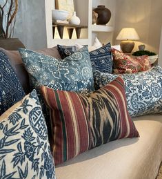 a couch with many pillows on it in front of a lamp and shelves filled with vases