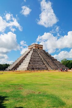 an ancient pyramid in the middle of a green field with people walking around it and clouds overhead