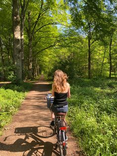 a woman riding a bike down a dirt road through a forest filled with green trees