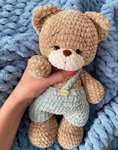 a hand holding a teddy bear on top of a blue knitted blanket with a tag in it's ear