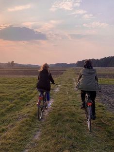 two people riding bikes down a dirt path in the middle of a field at sunset