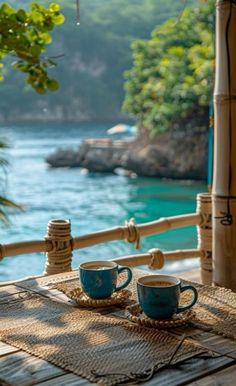 two coffee cups sitting on top of a wooden table next to the ocean with bamboo rafters