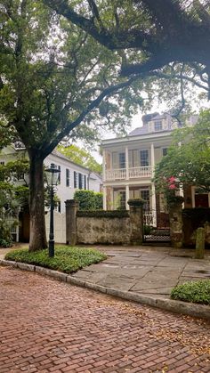 an old brick walkway leading to a white house with large trees in the front yard