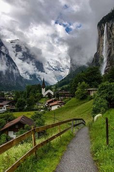 a path leading to a village with mountains in the back ground and clouds above it