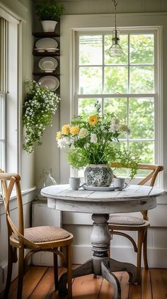 a white table with two chairs and flowers on it in front of a large window