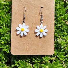 a pair of white and yellow flower earrings sitting on top of green plants next to a piece of brown paper