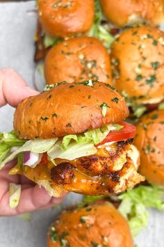 a hand is holding a sandwich with lettuce and tomato on it, while other sliders are in the background