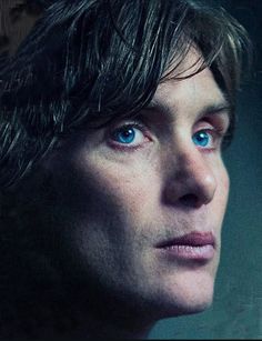 a man with long hair and blue eyes stares into the distance while standing in front of a dark background