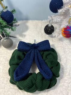 a green and blue knitted christmas wreath on a white blanket with ornaments in the background