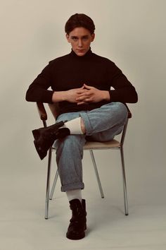 a man sitting in a chair with his hands folded and looking at the camera while wearing black boots